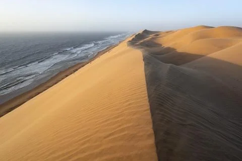 Sand dunes with wave patterns and sea in evening light, wind blowing sand across Stock Photos