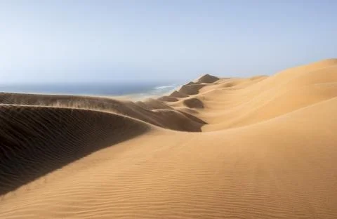 Sand dunes with wave patterns and sea in evening light, wind blowing sand across Stock Photos