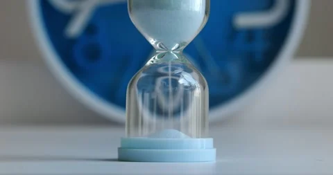 Sand flows through hourglass table before wall clock, moving second hand Stock Footage 267366480