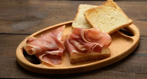 Sandwich with a white square slice of bread and slices of prosciutto on a woo Foto stock