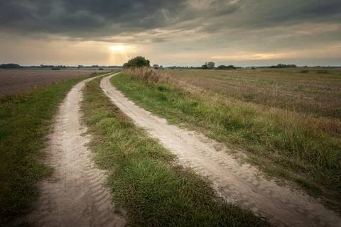 A sandy dirt road through fields and a cloudy evening sky Stock Photos