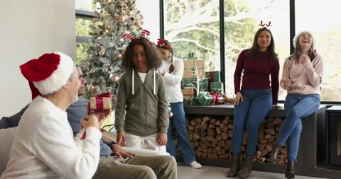 Santa-hat man handing gift to child on couch near Christmas tree, child untying Stock Footage 335453890