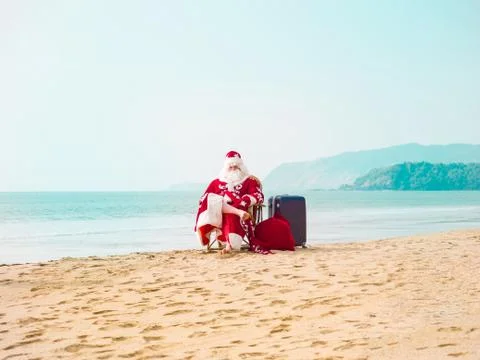 Santa with a suitcase and a bag with gifts sits on the beach on a journey Stock Photos
