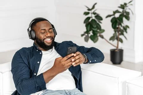 Satisfied handsome African guy using smartphone and wireless headphones Stock Photos
