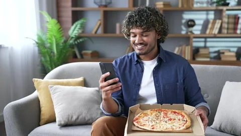 Satisfied young man holds an open box with pizza sitting on sofa in living room  Видео 334227047