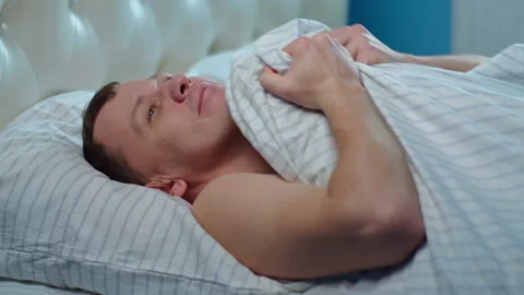 Satisfied young man lies down in bed, covers himself with a blanket Stock Footage 141536593
