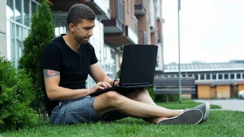 Satisfied young man plays computer game on a laptop while sitting on the grass Stock Footage 111974618