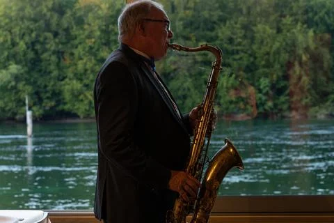 Saxophone player playing jazz in front of windows with forest and blue river Stock Photos