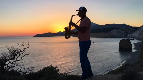 The saxophonist plays in the rays of the sunset on a cliff overlooking the sea Stock Footage 86561887