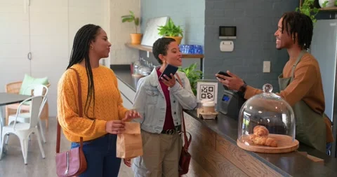 Scanning QR code to pay at counter, female friends receiving coffee cups on tray Vídeo Stock 337705012