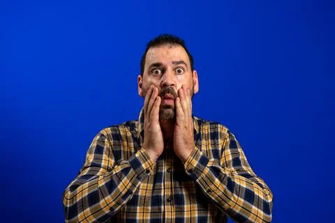Scared bearded man with his hands on his face and wide eyes isolated on blue  Stock Photos