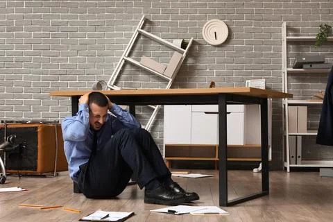 Scared businessman hiding under table in office during earthquake Stock Photos