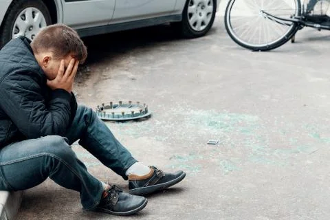 Scared driver sitting on a pavement next to his car and broken glass on the s 스톡 사진
