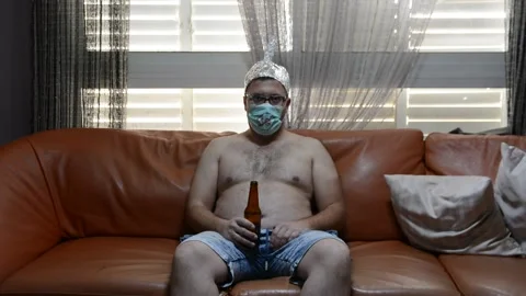 Scared freak man on a sofa in a foil hat and glasses drink beer. freak man with  Stock Footage 130879139