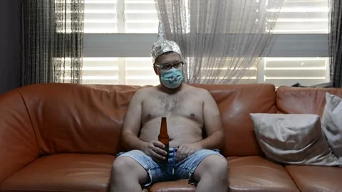 Scared freak man on a sofa in a foil hat and glasses drink beer. freak man with  Stock Footage 130879141