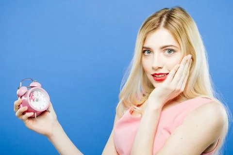 Scared Young Blonde is Looking at the Camera Holding Pink Alarm Clock in Her Stock Photos