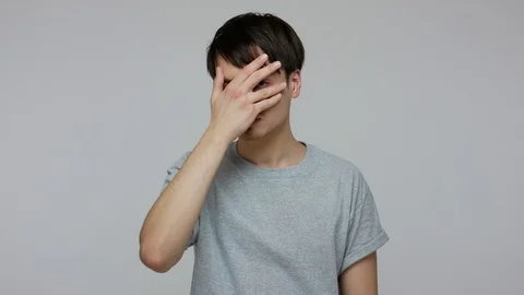 Scared young guy in T-shirt covering his face with hand, looking through fingers 스톡 동영상 124772986