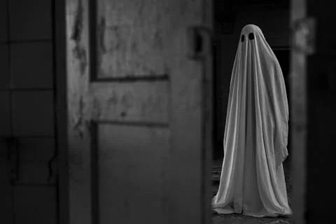 Scary ghost covering in white sheet on dark background near open door in aban Stock Photos