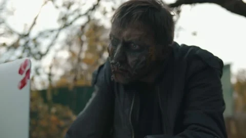 Scary zombie using laptop sitting outdoors in abandoned courtyard Stock Footage 141192956