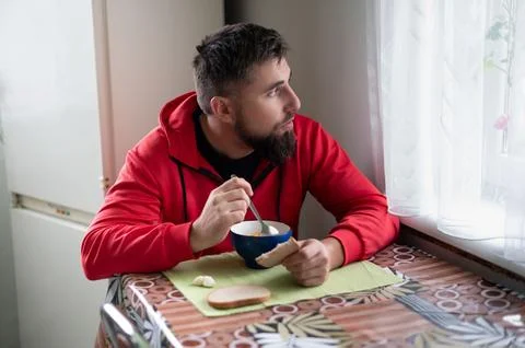 This scene captures a bearded man in a red hoodie eating soup at a patterned  Foto stock