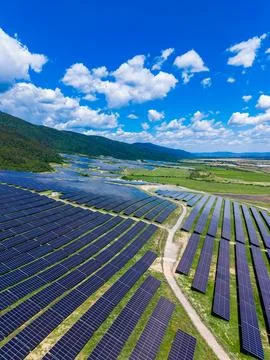 The scene shows rows of solar panels set in green fields. Mountains are in th Stock Photos