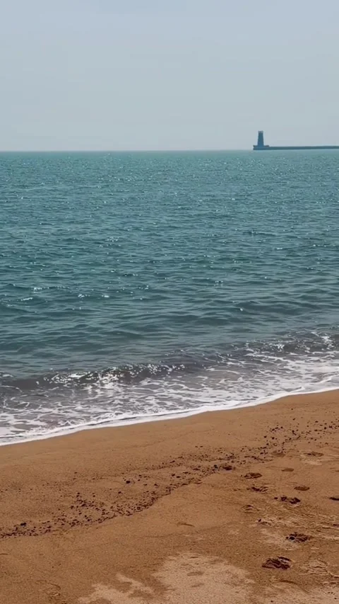 Scenic view of a sandy beach with gentle waves and a distant lighthouse on the Vidéo 334163151