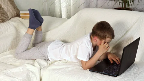 School boy lying on cozy sofa and cratching his forehead studying with laptop Stock Footage 154234264
