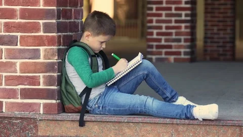 School boy sitting in the schoolyard, doing homework and waits for parents Stock Footage 156078624