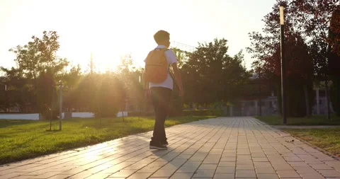 Schoolboy with backpack walks along paved park path at sunset. Student enjoys Stock Footage 345477256