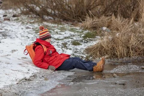A schoolboy boy with a backpack wearing a red coat and a striped hat sits o.. Stock Photos