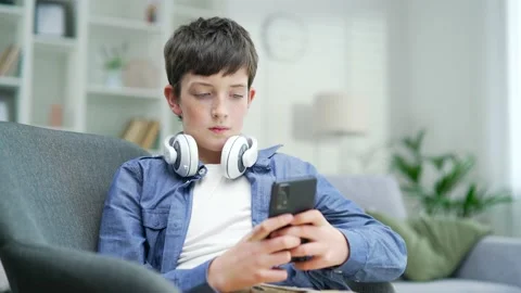 A schoolboy boy uses a smartphone while sitting on the couch at home. Indoor.  Stock Footage 238633270