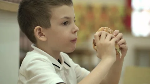 Schoolboy eats hamburger sitting in cafe. Boy holds a burger in his hands and ea Stock Footage 219579823