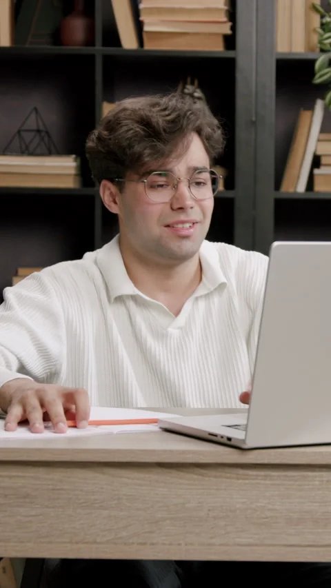 Schoolboy with glasses sits at desk, focused on laptop screen, surrounded by Video stock 332473596
