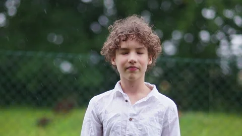 Schoolboy has fun in the warm summer rain. Close portrait curly boy in white Stock Footage 133723531