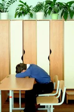 Schoolboy with head down on desk in classroom, surrounded by wooden lockers and Stock Photos
