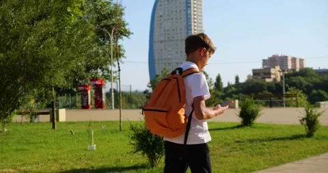 Schoolboy with orange backpack checks smartphone in city park. Young student Stock Footage 346042296
