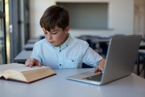 Schoolboy reading a book and using laptop in a classroom Stock Photos