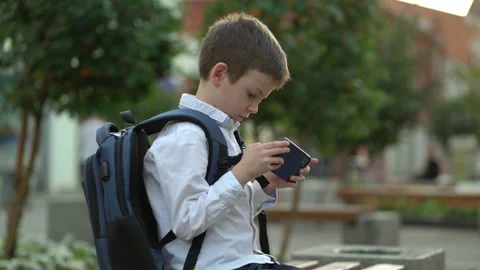 A schoolboy sits on a bench in the park and plays on his phone after studying at Stock Footage 204882941