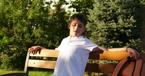 Schoolboy sits on bench in sunny park with backpack. Boy relaxes outdoors after Stock Footage 345475718