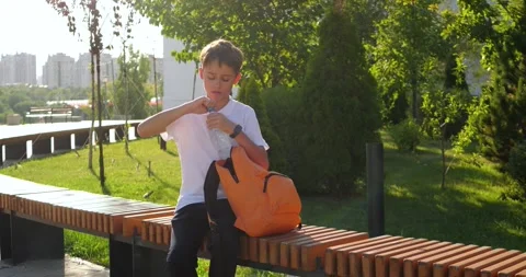 Schoolboy sits on bench in sunny park, takes water bottle from orange backpack Stock Footage 346041693