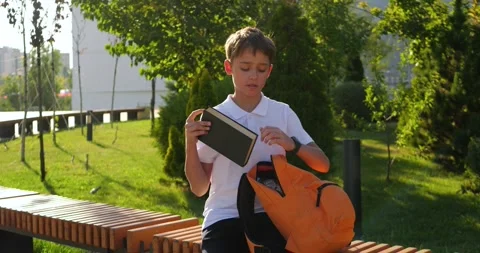 Schoolboy sits on park bench and opens orange backpack. Boy takes out book and Stock Footage 346691402
