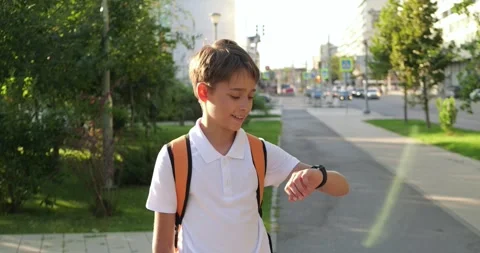 Schoolboy stands on city sidewalk with backpack, checks wristwatch and smiles Stock Footage 345477089