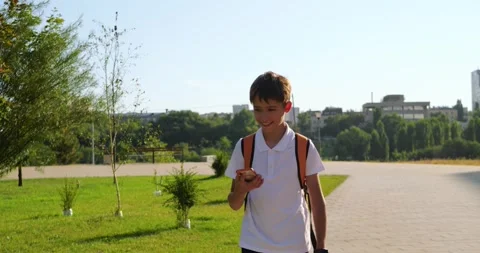 Schoolboy walks through city park with backpack, takes smartphone from pocket Stock Footage 345475959