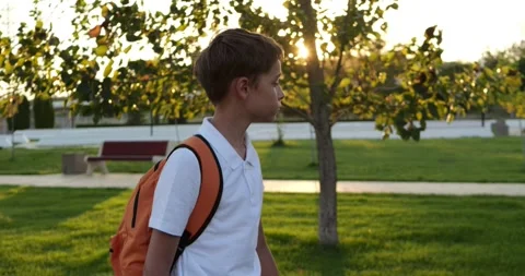 Schoolboy walks through park carrying orange backpack. Young student heads home Stock Footage 345787566