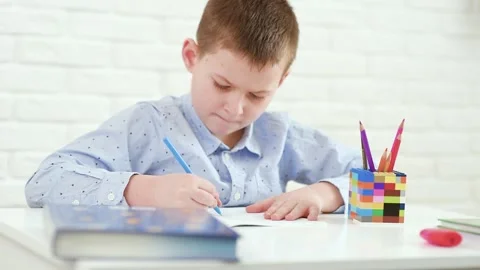Schoolboy writes in a notebook sitting at a white table. Stock-Footage 151382409