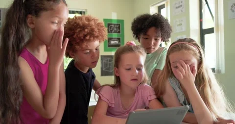 Schoolchildren leaning around tablet at classroom table, central girl touching Stock Footage 341320338