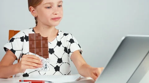 Schoolgirl eating chocolate and typing in her laptop sitting at the desk Stock Footage