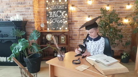 A schoolgirl in a graduate cap is studying a microscope, sitting at her desk Stock Footage 169048003