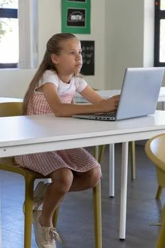 Schoolgirl sitting at table using silver laptop in class wearing pink pinafore Stock Photos