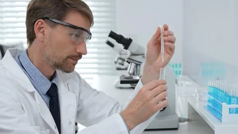Scientist in lab coat and protective glasses adding reagent to test tube with Stock Photos
